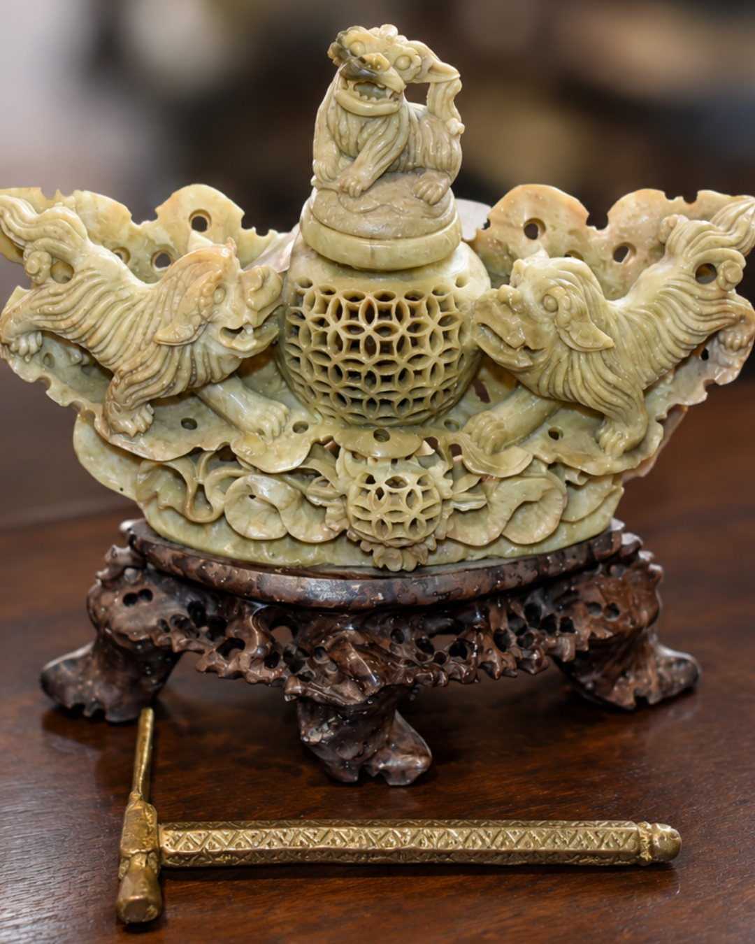 An ornate carved jade decorative object with a small figurine on top, set on a dark wooden stand, on a wooden surface.