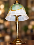 An ornate table lamp with a textured gold metal base and a bell-shaped glass shade featuring a white lower section and a green upper section with gold detailing, set against a warm, blurred background.