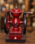 An antique red and black hand-crank coffee grinder with a wooden handle and base, displayed on a wooden table with a warm, blurred background.