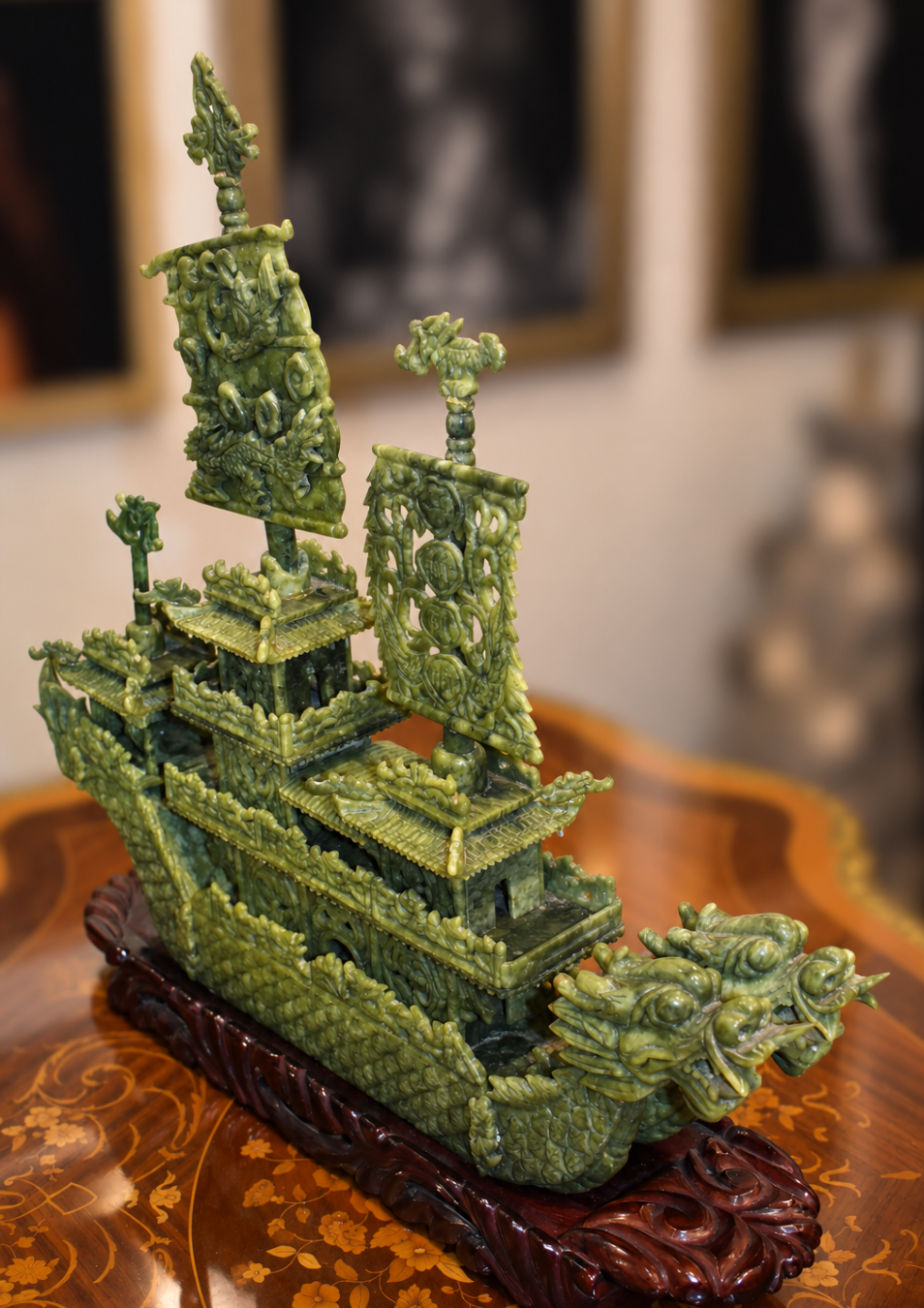 An intricately carved green jade ship sculpture displayed on a dark wooden base, set against a softly blurred indoor background.