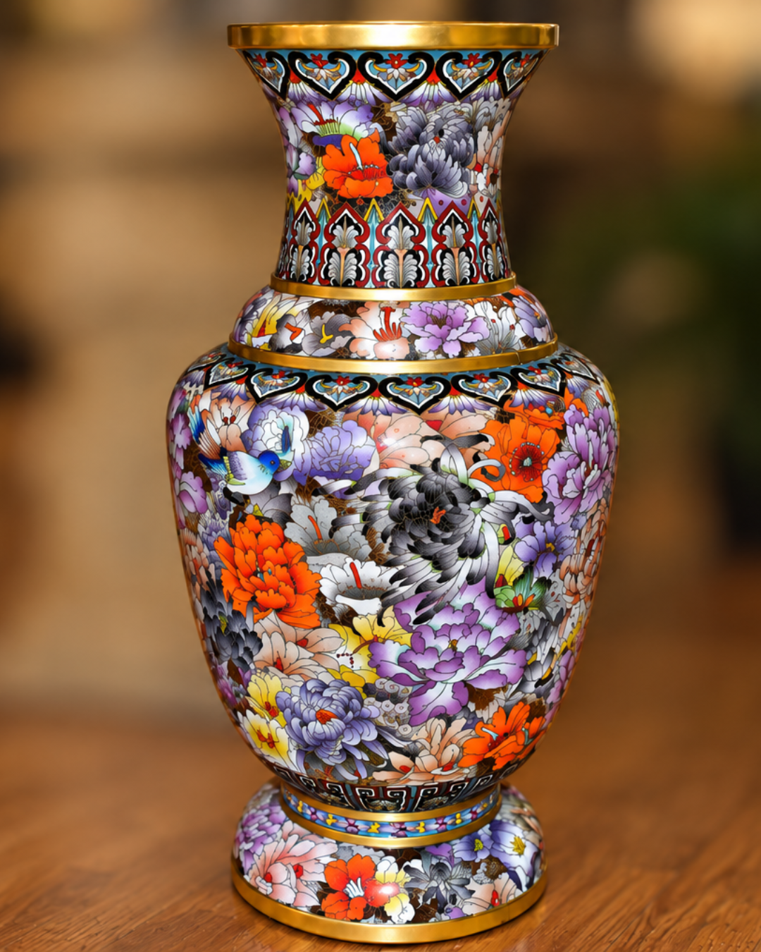 A decorative ceramic vase with a highly detailed, colorful floral pattern on a dark background, featuring gold trim at the neck, base, and rim, set on a wooden surface with a blurred background.