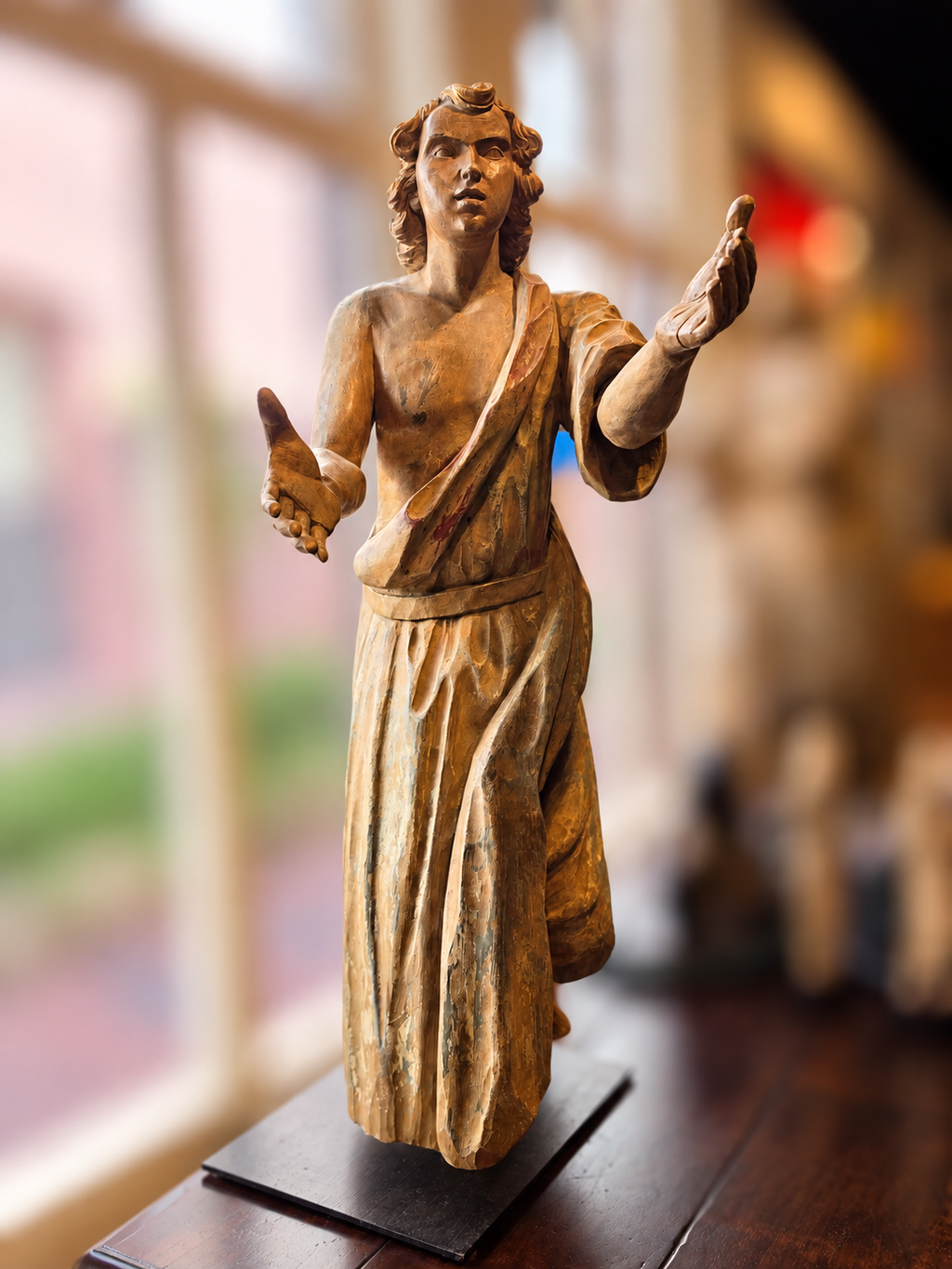 A carved wooden statue of a robed man with outstretched hands, displayed on a dark base in a softly blurred indoor setting.