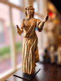 A carved wooden statue of a robed man with outstretched hands, displayed on a dark base in a softly blurred indoor setting.