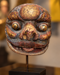 Front view of a painted Asian cultural mask with rounded eyes, arched brows, and an open mouth, mounted on a black display stand
