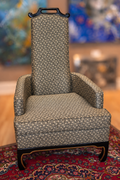 A tall upholstered armchair with a geometric patterned fabric, dark wood frame, and high backrest, shown standing on a red patterned rug in an indoor room with blurred artwork in the background.