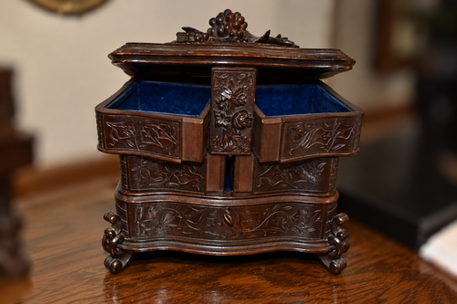 An ornate dark brown carved wooden jewelry box with a hinged lid, blue velvet lining, and decorative floral engravings, shown on a wooden table in a softly blurred indoor setting.