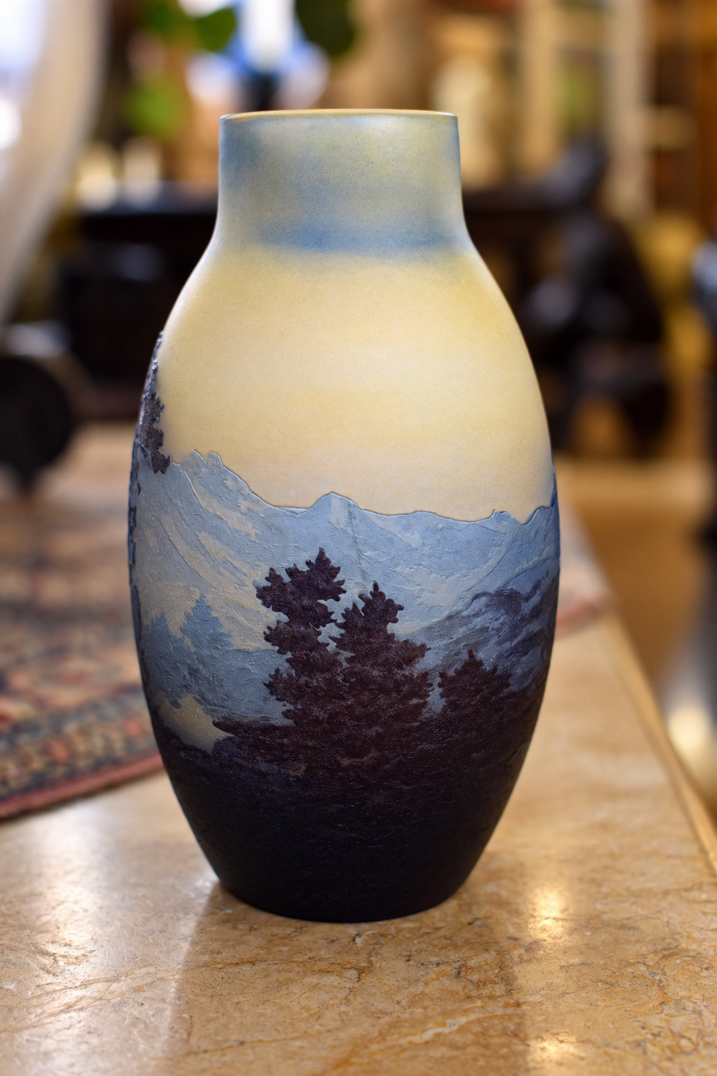 A ceramic vase with a hand-painted mountain landscape design, featuring blue and purple mountains, dark pine trees, and a cream-to-light-blue gradient sky, displayed on a marble countertop.