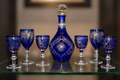 Cobalt blue cut glass decanter with a clear stopper, flanked by six matching stemmed glasses on a glass shelf.