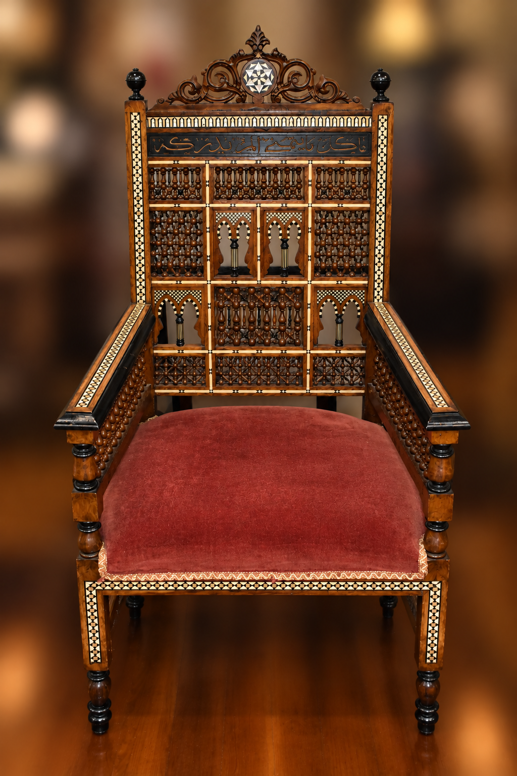 Front view of a Middle Eastern wood armchair with mother-of-pearl inlay, an intricately carved back, open arms, and a red upholstered seat