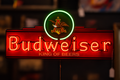 A glowing Budweiser neon sign with a red illuminated background, the Budweiser name in large red letters, the Budweiser crest above, and the words 'King of Beers' below, photographed against a dark blurred bar-like background.