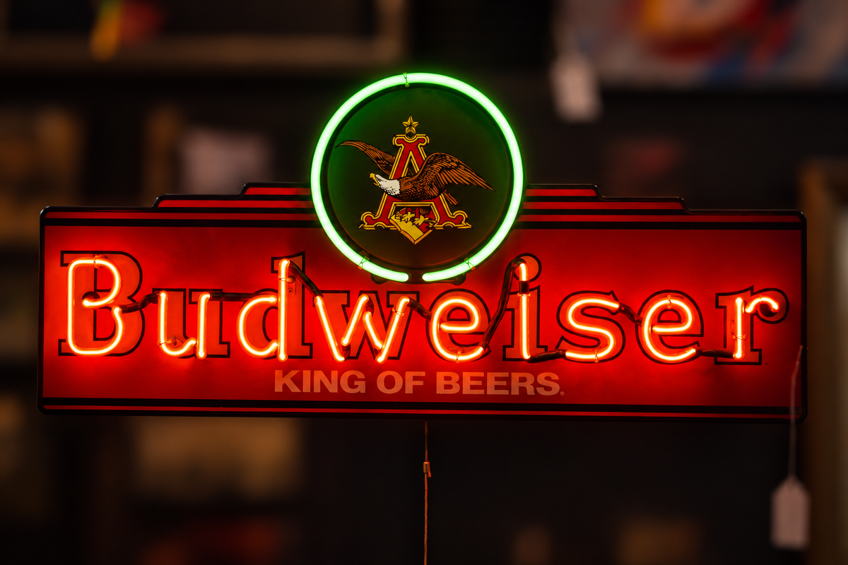 A glowing Budweiser neon sign with a red illuminated background, the Budweiser name in large red letters, the Budweiser crest above, and the words 'King of Beers' below, photographed against a dark blurred bar-like background.