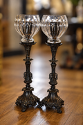 A pair of ornate black metal candlestick holders with clear cut-glass globes, displayed on a polished wooden floor with a softly blurred indoor background.