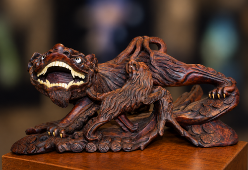 A detailed hand-carved wooden sculpture of a mythical beast with a wide-open mouth showing sharp teeth, perched on a cluster of lotus buds, displayed on a wooden surface with a blurred background.