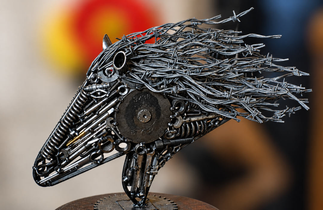 A detailed close-up of a metal sculpture depicting a bird's head and neck, constructed from various metallic parts, springs, gears, and barbed wire, mounted on a round wooden base.