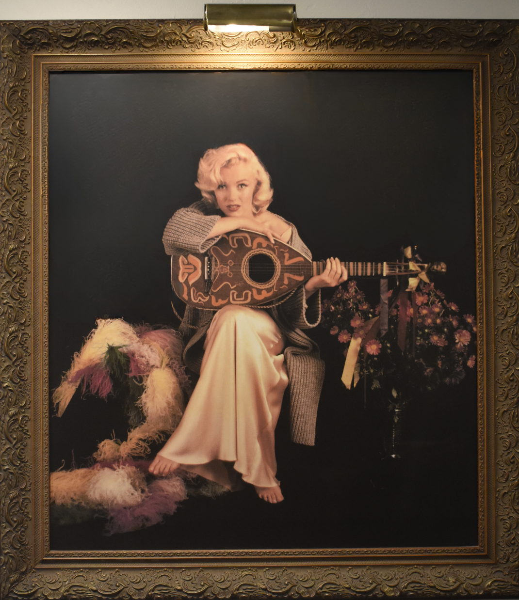 A large framed portrait print depicting Marilyn Monroe seated and holding a decorative guitar against a dark background, enclosed in an ornate gold frame. The artwork is photographed hanging on a wall under a light fixture.