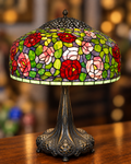 A Tiffany-style table lamp with a stained-glass shade featuring red, pink, and white roses with green leaves, mounted on an ornate dark metal stem and base, set on a wooden surface with a blurred warm background.