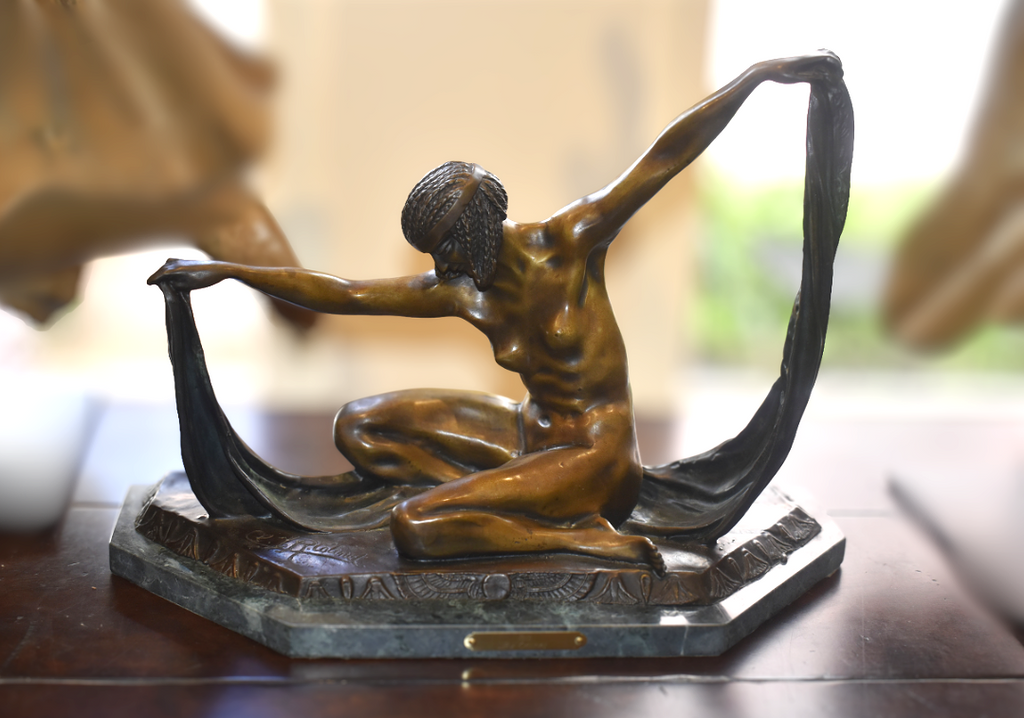 A bronze sculpture of a kneeling nude female figure with arms raised, holding a long dark drapery shape, displayed on a dark wooden surface with a blurred indoor background.