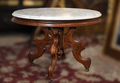 An antique round pedestal table featuring a white marble top and an ornate carved dark brown wooden base with casters, set on a patterned red rug.