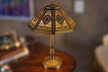 An ornate vintage-style table lamp with a gold and black geometric patterned shade and a polished brass base, displayed on a wooden side table in a dimly lit room.