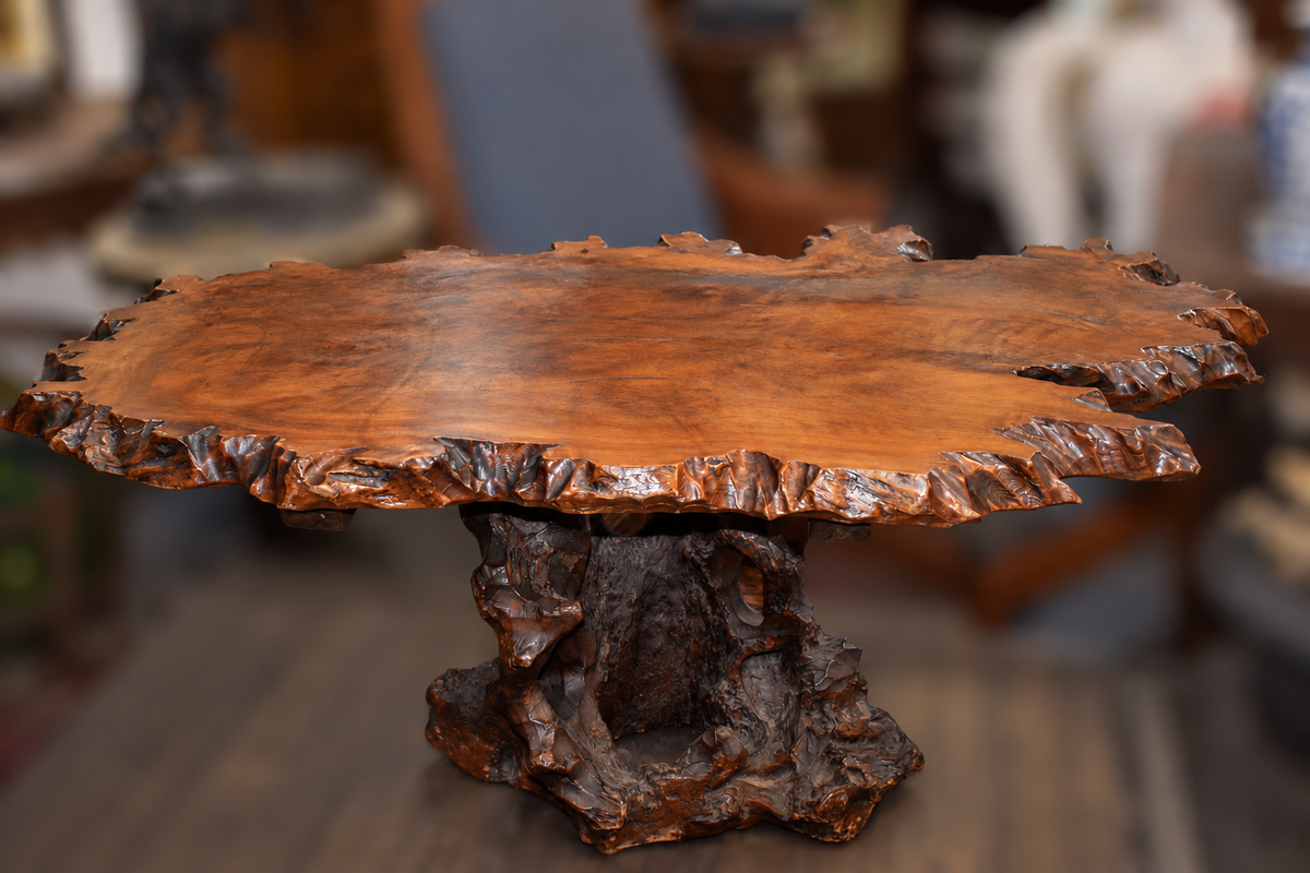 A rustic live-edge wooden table with a richly textured, polished brown wood top and a sculptural, root-like dark brown pedestal base, shown in a softly blurred indoor setting.