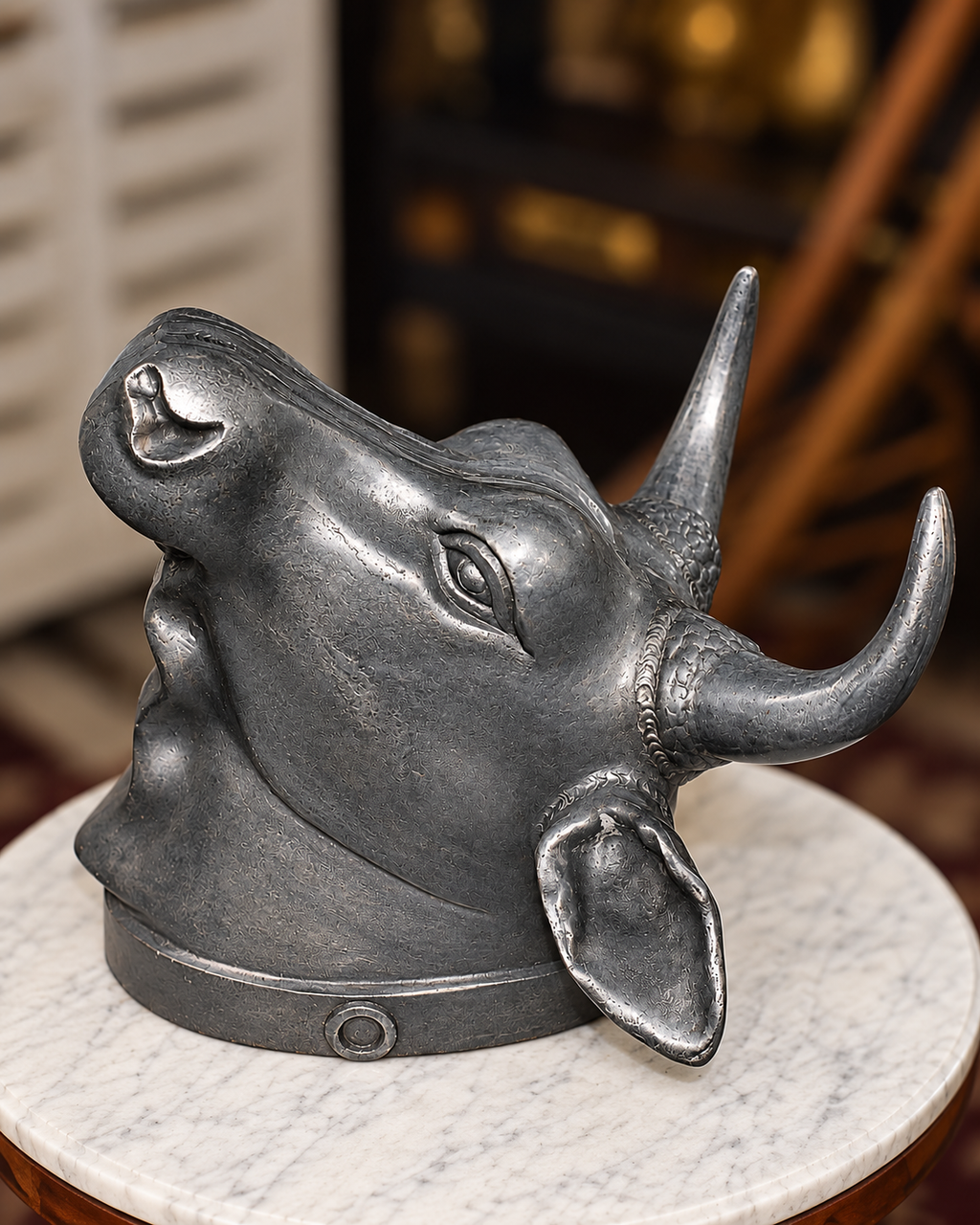 A dark metallic bull head sculpture or decorative mask with prominent horns, displayed on a round white marble pedestal in an indoor setting with a blurred background.