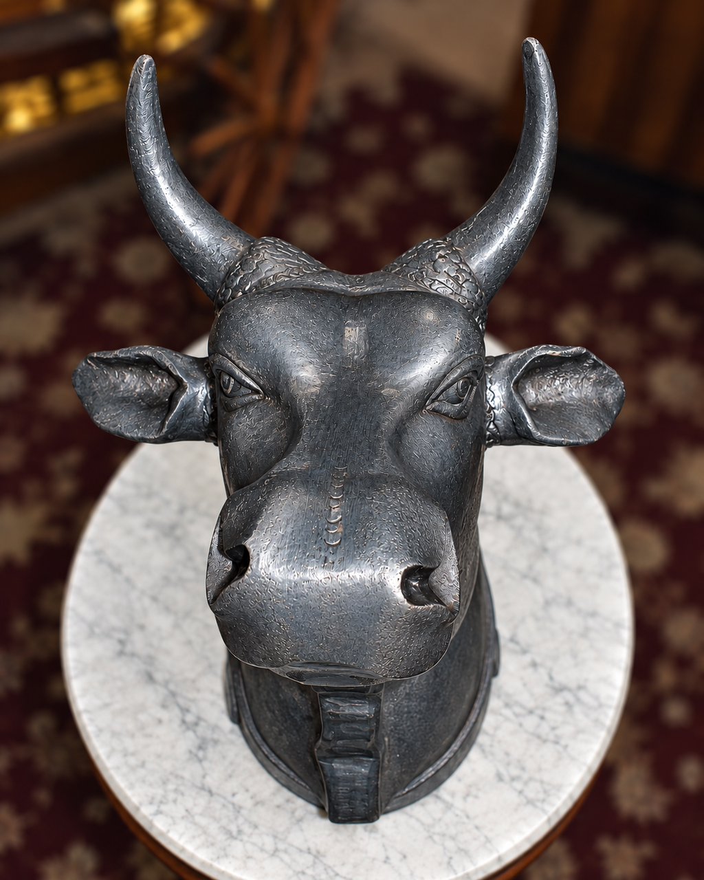 A dark metallic bull head sculpture with curved horns, mounted on a round white marble base in an indoor setting with a patterned rug in the background.