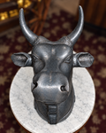 A dark metallic bull head sculpture with curved horns, mounted on a round white marble base in an indoor setting with a patterned rug in the background.